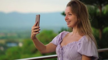 Arka planda güzel doğa manzarası olan bir kadın selfie çekiyor. Gülümseyen kadın gündelik yazlık elbiseler giyiyor ve modern teknolojiyle bağlantının tadını çıkarıyor. Açık hava eğlencesi ve dijital
