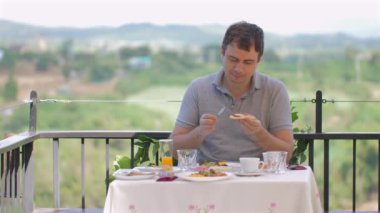 Orta yaşlı bir adam arka planda manzaralı balkonda kahvaltıdan hoşlanıyor. Masa meyve suyu, kahve ve hamur işleri de dahil olmak üzere çeşitli kahvaltı malzemeleriyle hazırlandı.