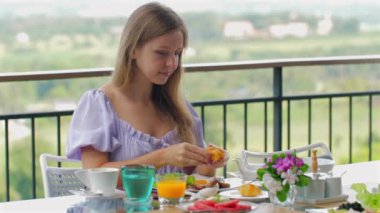 Genç bir kadın güneşli balkonda manzaralı sağlıklı bir kahvaltı yapıyor. Taze meyve, meyve suyu, kahve ve hamur işleri güzel bir sehpada. Yeşillik ve çiçekli manzara, gevşemiş sabahı destekliyor.