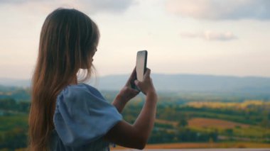 Genç bir kadın akıllı telefonuyla gün batımında yuvarlanan tepelerle ve resimli gökyüzüyle manzaralı bir yerde fotoğraf çekiyor. Turizm ve teknoloji.