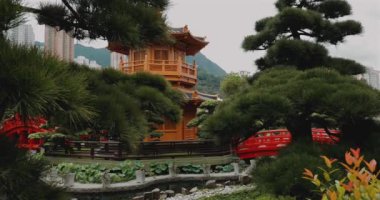 Geleneksel Japon bahçesi ahşap pagoda, canlı kırmızı köprü, yemyeşil ve arka planda şehir manzarası. Doğayı mimariyle harmanlayan huzurlu bir manzara. Açık hava zen bahçesi sahnesi.