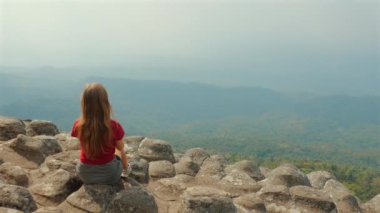 Kırmızı tişörtlü, uzun saçlı bir kadın kayalık kayalıklarda oturuyor ve engin bir dağ manzarasına bakıyor. Yalnızlık ve Doğa Takdir..