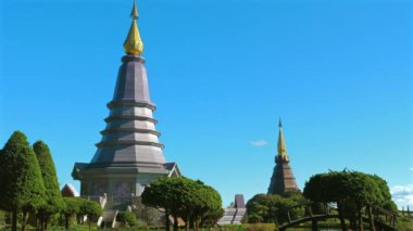 Parlak mavi gökyüzü altındaki yemyeşil bahçelerle çevrili iki büyüleyici Tayland pagodasının manzarası. Geleneksel mimari ve canlı yeşillik. Kültür Mirası ve Seyahat.
