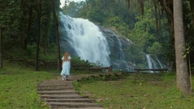 Young woman in long dress walking towards majestic waterfall in lush forest, natural beauty and tranquility inspired by nature, Adventure and Relaxation.