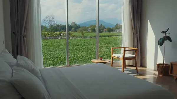 Modern minimalist yatak odası panoramik pencere manzaralı yemyeşil tarlalar ve dağlar, rahat ahşap sandalye ve saksı bitkisi, sükunet ve doğal güzellik. Rahatlama ve Dinginlik.