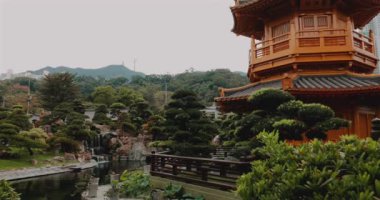Geleneksel Japon bahçesi, yemyeşil, küçük ağaçlar, dingin gölet, ve detaylı ahşap pavyon. Sakin gökyüzü altında sakin bir dağ arkaplanı. Barış, doğa ve Asya kültürü teması için ideal..