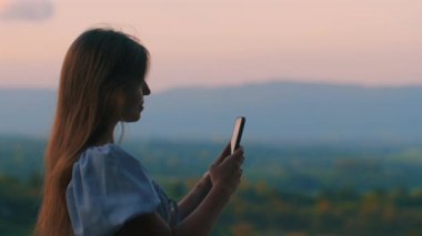 Gün batımında uzun saçlı genç bir kadın akıllı telefona bakıyor. Arka planda tepelerin ve ormanın resimli manzarası olan sakin bir açık hava manzarası. Teknoloji ve doğadaki iletişim