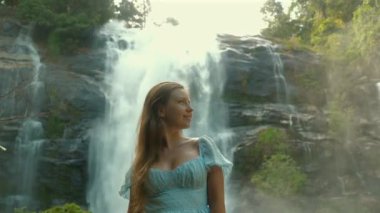 Mavi elbiseli genç kadın gür ormanlarda görkemli şelalenin önünde duruyor, doğanın ve sükunetin kıymetini biliyor. Seyahat ve Macera.