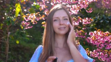 Çiçek açan kiraz ağaçları altında cilt bakımı ürünleri uygulayan genç bir kadın doğal güzelliği ve kendine bakımı vurguluyor. Doğal Cilt Bakımı ve Kendine Bakım