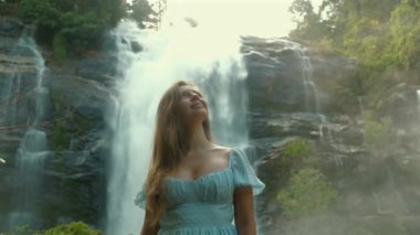 Görkemli şelalede dingin anın tadını çıkaran bir kadın. Mavi elbise giymiş, yemyeşil bir manzarayla çevrili. Doğa ve Sağlık