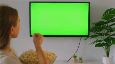 Genç kadın evde yeşil ekran televizyon izlerken patlamış mısırdan zevk alıyor, rahat oturma odası, eğlence ve gevşeme..