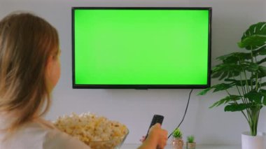 Kadın elinde patlamış mısır kasesi ve uzaktan kumandayla modern televizyonda boş yeşil ekran seyrediyor. Sıcak ve rahatlatıcı bir eğlence ortamı. - Ev Eğlencesi ve Rahatlama