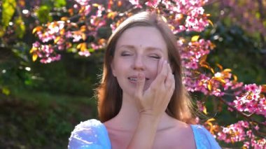 Kadın nazikçe yüz kremi sürüyor, bahçede çiçek açan kiraz çiçeklerinin yanında duruyor, doğanın ve baharın canlı renklerinin tadını çıkarıyor. Rahatlama ve Doğal Güzellik