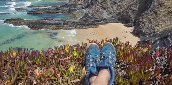 Trekker feet resting on the edge of the cliff. Cabo Sardao coast, Ponta do Cavaleiro, Sao Teotonio, Portugal