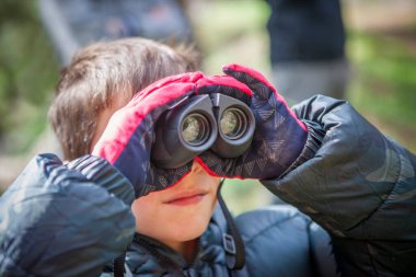 Çocuk doğada dürbün tutuyor. Eldiven ve kamuflaj anorak giyiyor.