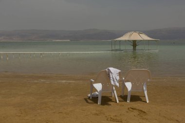 Ölü Deniz 'de güneşlik ve kumlu sahilde havluyla beyaz plastik sandalyeler.