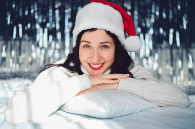 Kırmızı rujlu, beyaz kazaklı, güzel, gülümseyen esmer kadın portresi, Noel Baba şapkası evde, mutlu Noeller. Yeni yıla ışıldayan ışıkları olan neşeli bayan.