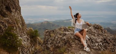 Genç kadın serbest gezgin dışarıda herhangi bir yerde dizüstü bilgisayarla çalışarak dağ manzarasının tadını çıkarıyor. Mutlu kadın vites küçültücü yazın olağandışı manzaralı iş yerinde bilgisayar tutuyor.