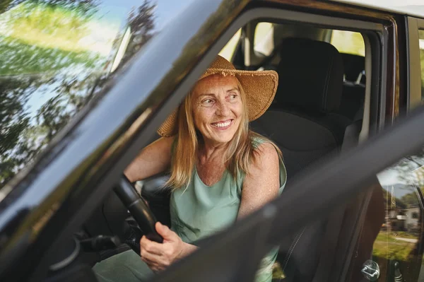 Happy senior woman driver in straw hat driving sitting in new car, smiling looking at camera enjoying journey. Driving courses and life insurance. Retired people activity and road trip concept