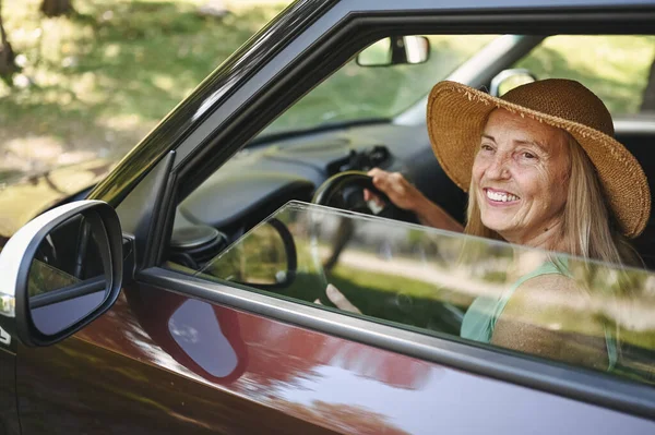 Happy senior woman driver in straw hat driving sitting in new car, smiling looking at camera enjoying journey. Driving courses and life insurance. Retired people activity and road trip concept