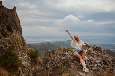Genç kadın serbest gezgin dışarıda herhangi bir yerde dizüstü bilgisayarla çalışarak dağ manzarasının tadını çıkarıyor. Mutlu kadın vites küçültücü yazın olağandışı manzaralı iş yerinde bilgisayar tutuyor.