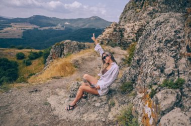 Genç kadın serbest gezgin dışarıda herhangi bir yerde dizüstü bilgisayarla çalışarak dağ manzarasının tadını çıkarıyor. Güneş gözlüklü mutlu kadın şekil değiştiren yazın sıra dışı bir iş yerinde bilgisayar tutuyor.