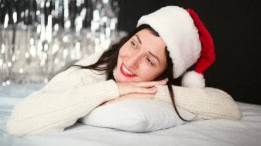Kırmızı rujlu, beyaz kazaklı, güzel, gülümseyen esmer kadın portresi, Noel Baba şapkası evde, mutlu Noeller. Yeni yıla ışıldayan ışıkları olan neşeli bayan.