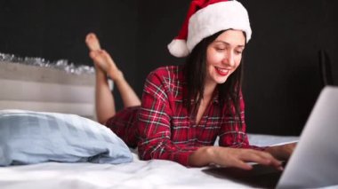 Kırmızı pijamalı güzel, heyecanlı esmer kadın Noel Baba şapkasıyla yatakta uzanıyor. İç odasındaki karanlık dizüstü bilgisayarla çalışıyor. Genç bayan serbest çalışan mutlu Noel havasında evde..