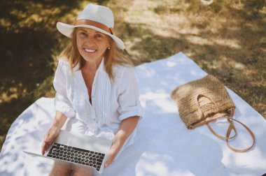 Kıdemli mutlu kadın çalışıyor ya da yazın dışarıda dizüstü bilgisayarla görüntülü görüşme yapıyor. Hasır şapkalı, çantalı, kırsal kesimdeki piknik sırasında çimlerin üzerinde oturan zarif bir hanım. Aktif emekliler konsepti