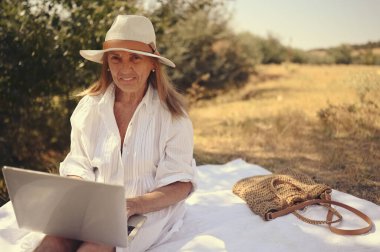 Kıdemli mutlu kadın çalışıyor ya da yazın dışarıda dizüstü bilgisayarla görüntülü görüşme yapıyor. Hasır şapkalı, çantalı, kırsal kesimdeki piknik sırasında çimlerin üzerinde oturan zarif bir hanım. Aktif emekliler konsepti