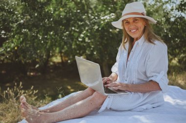 Kıdemli mutlu kadın çalışıyor ya da yazın dışarıda dizüstü bilgisayarla görüntülü görüşme yapıyor. Hasır şapkalı, çantalı, kırsal kesimdeki piknik sırasında çimlerin üzerinde oturan zarif bir hanım. Aktif emekliler konsepti
