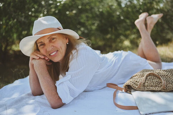 Senior happy woman laying on picnic blanket with laptop in bag at summer outdoors. Old elegant lady in straw hat on grass at countryside. Active retired people concept