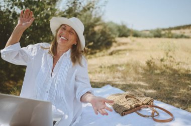 Kıdemli mutlu kadın çalışıyor ya da yazın dışarıda dizüstü bilgisayarla görüntülü görüşme yapıyor. Hasır şapkalı, çantalı, kırsal kesimdeki piknik sırasında çimlerin üzerinde oturan zarif bir hanım. Aktif emekliler konsepti