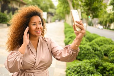 Çekici mutlu stil artı Afrikalı Amerikalı kadın öğrenci serbest afro saç dışarıda akıllı telefonla selfie çekiyor. Yazın büyük şehir caddesinde ipek elbiseli genç bayan. Çeşitlilik. 
