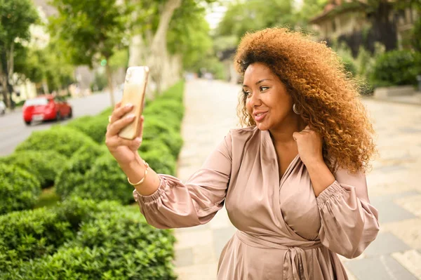 迷人的快乐时尚加尺寸的非洲裔美国女学生自由职业者用智能手机自己做头发 夏天穿着丝绸衣服的年轻女士住在大城市街道上 多样性 图库图片