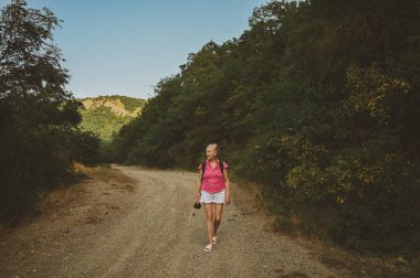 Gün batımında yaz ormanı yolunda gezen mutlu, son sınıf öğrencisi bir turist. Sıska kadın fotoğraf makinesiyle seyahat ediyor. Aktif emeklilik tatili konsepti. Sıcak filtre