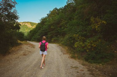 Kıdemli kadın sırt çantalı turist yaz ormanı yolunda gün batımında arka planda yürüyor. Yaşlı sıska bayan yalnız seyahat ediyor. Aktif emeklilik tatili ve sağlıklı yaşam tarzı konsepti. Sıcak filtre.