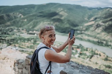 Olgun, mutlu, gülümseyen bir turist yaz tatilinde gözlem güvertesinde fotoğraf çeker. Emekli insanlar yaz tatili, aktif yaşam tarzı konsepti. 