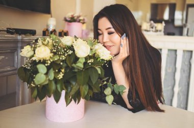 Güzel Asyalı kadın oturma odasındaki masada oturuyor parlak iç mekan beyaz güllerin çiçek buketine hayran. Gülümseyen genç bayan sürpriz bir hediye almaktan mutluluk duyar.