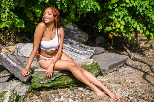 Açık hava portresi. Kırmızı örgü saç stili ve mükemmel beyaz dişli güzel Afro-Amerikan kadın. Güneşli yaz gününde, plaj arka planında kayalarla mayo giymiş.