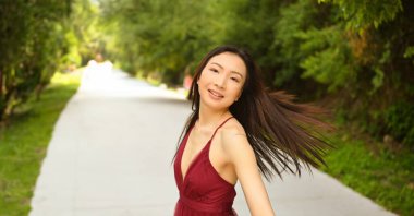Güneşli yaz gününün dışında yürüyen güzel, genç, Asyalı mutlu bir kadının sokak tarzı portresi. Kırmızı elbiseli çekici bir kadın yeşil parkta poz vermekten hoşlanıyor.