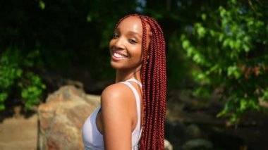 Doğal afro-amerikalı güzel bir kadının açık hava portresi uzun kırmızı örgü saç stili ve mükemmel beyaz diş tebessümü, güneşli yaz gününde yeşil yeşillik arka planıyla mayo giymiş.