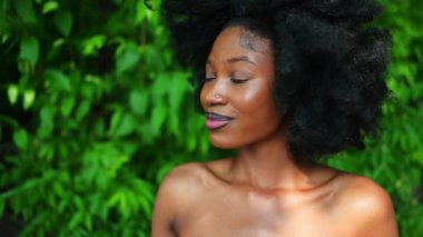 Yavaş çekimde, güzel Afro-Amerikan kadın mükemmel beyaz dişler dışarıda gülümsüyor, güneşli yaz günü yeşil yapraklar. Yüz bakımı. Kozmetoloji, cilt bakımı ve kaplıca