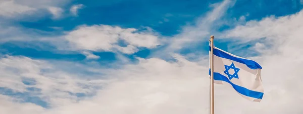 Üzerinde Davut Yıldızı olan İsrail bayrağı bulutlu mavi gökyüzüne doğru sallanıyor, panoramik bir manzara. Ulusal devlet sembolleri ve taklit uzay afişleriyle vatansever İsrail kavramı