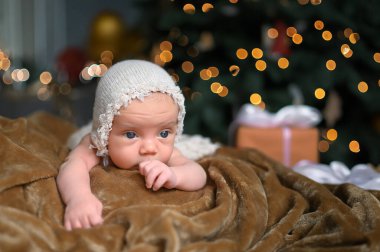 Yeni yıl tatili sahnesi. Yeni doğan komik şapkalı, bej bir battaniyenin üzerine uzanmış şirin bir bebek. Noel ağacı oyuncakları ve ışık çelengi. Bebek eşyaları konsepti, kreş