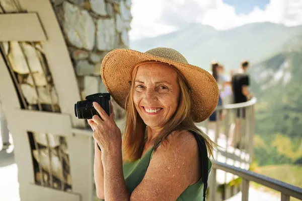 Portrait of a smiling happy elderly woman tourist traveling with a camera posing against the backdrop of mountains. Old elegant lady in straw hat on grass at countryside. Active retired people concept