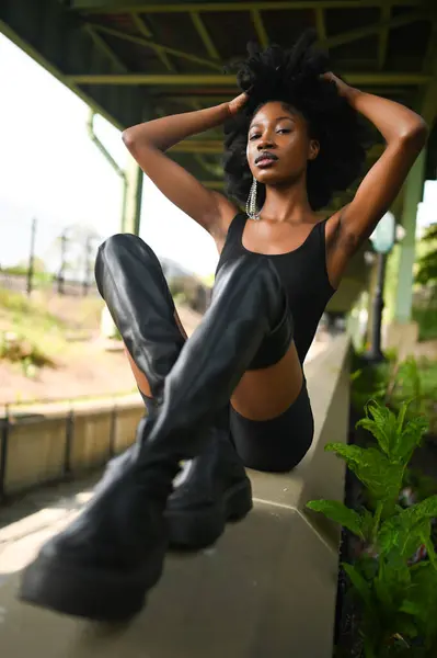 Fashion outdoor street style portrait Beautiful young African American woman posing outside on urban city landscape summer day wearing total black high leather boots.