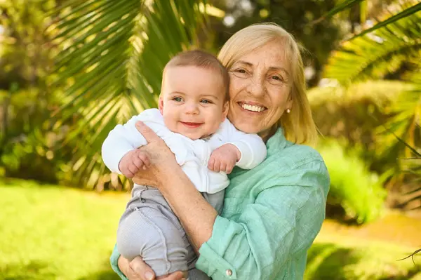 Beautiful happy smiling senior elderly woman holding on hands cute little baby boy. Grandmother and grandson having fun time together at tropical summer day in park. Multigenerational family concept