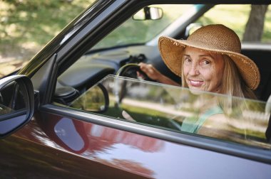 Hasır şapkalı, yeni arabasında oturan, gülümseyen ve kameraya bakıp yolculuğun tadını çıkaran son sınıf öğrencisi mutlu kadın sürücü. Sürücü kursları ve hayat sigortası. Emekli insanlar etkinlik ve yolculuk konsepti 