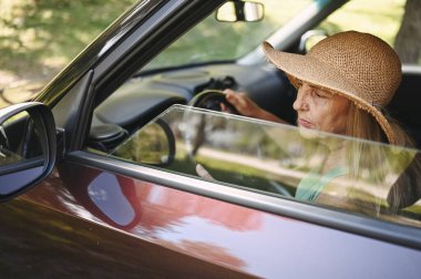 Hasır şapkalı, yeni arabasında oturan, gülümseyen ve kameraya bakıp yolculuğun tadını çıkaran son sınıf öğrencisi mutlu kadın sürücü. Sürücü kursları ve hayat sigortası. Emekli insanlar etkinlik ve yolculuk konsepti 
